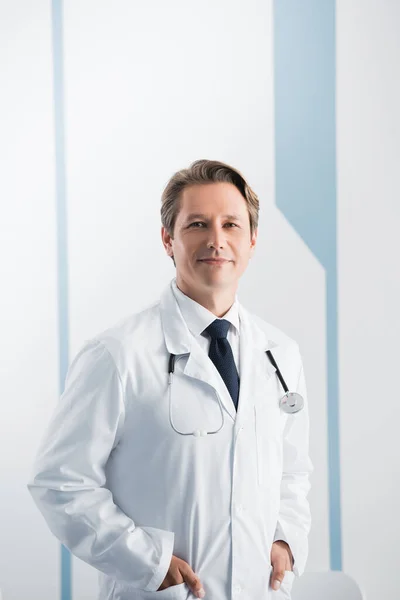 Doctor with stethoscope and hands in pockets looking at camera in hospital — Stock Photo