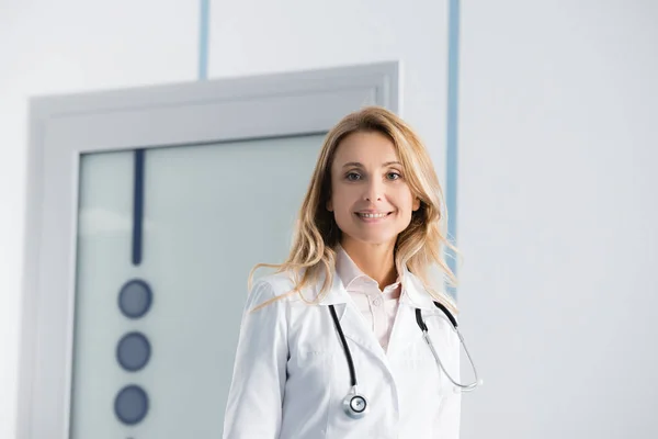 Médico rubio en bata blanca mirando a la cámara en el hospital - foto de stock