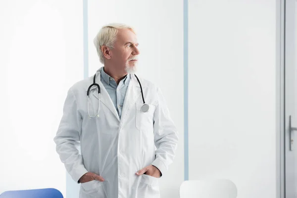 Médico de edad avanzada con bata blanca mirando hacia otro lado en el hospital - foto de stock