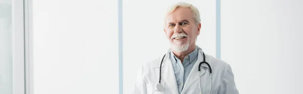Panoramaaufnahme eines betagten Arztes mit Stethoskop beim Blick in die Kamera in der Klinik — Stockfoto