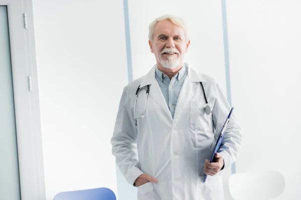 Grey haired doctor with hand in pocket holding clipboard in hospital — Stock Photo