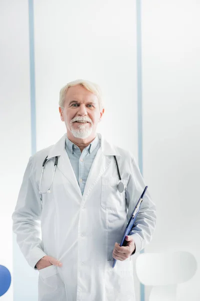 Elderly doctor with clipboard looking at camera in hospital — Stock Photo