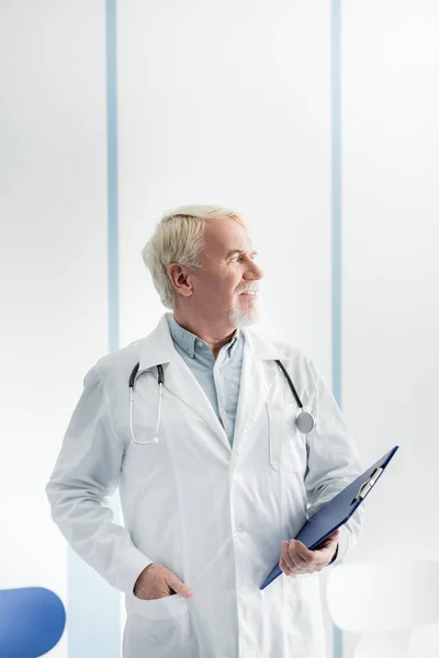 Grey haired doctor in white coat holding clipboard in hospital — Stock Photo