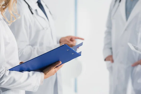 Cropped view of doctor holding clipboard near colleagues in clinic — Stock Photo
