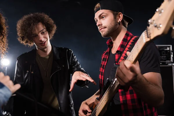 Curly young adult man pointing with hand at guitar, while standing near musician on black — Stock Photo