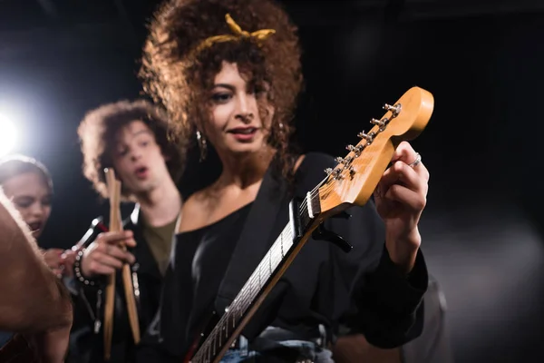 Curly woman touching and looking at electric guitar, while standing near musician pointing with finger on blurred background — Stock Photo