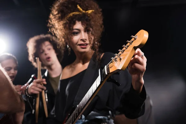 Femme bouclée touchant et regardant la guitare électrique, tout en se tenant près du musicien pointant du doigt sur fond flou — Photo de stock