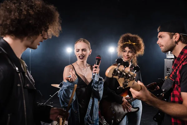 KYIV, UCRANIA - 25 de agosto de 2020: Mujer rubia escéptica haciendo gestos y mirando los muslos durante el ensayo de la banda de rock en un primer plano borroso - foto de stock