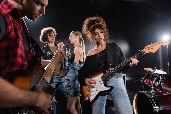 KYIV, UKRAINE - AUGUST 25, 2020: Guitarists playing electric guitars near talking rock band musicians with backlit and drum kit on black background — Stock Photo