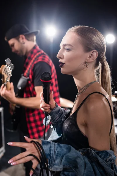 KYIV, UCRANIA - 25 de agosto de 2020: Mujer rubia haciendo gestos mientras sostiene el micrófono, de pie cerca del guitarrista sobre un fondo borroso - foto de stock