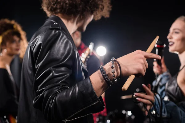 Curly man with drumstick pointing with finger during rehearsal with blurred rock band musicians on background — Stock Photo
