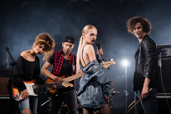 KYIV, UCRAINA - 25 AGOSTO 2020: musicisti rock band con chitarre elettriche, bacchette e microfono che guardano la fotocamera con retroilluminazione su nero — Foto stock