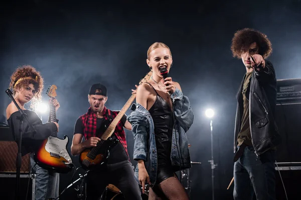 KYIV, UCRANIA - 25 de agosto de 2020: Mujer rubia cantando en el micrófono de pie cerca de músico señalando con la baqueta y guitarristas con retroiluminación en negro - foto de stock