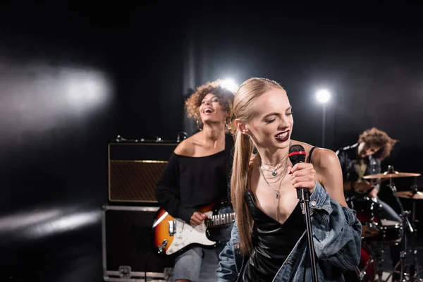 KYIV, UCRANIA - 25 de agosto de 2020: Mujer rubia cantando con micrófono de pie cerca del guitarrista con batería retroiluminada y borrosa en el fondo - foto de stock