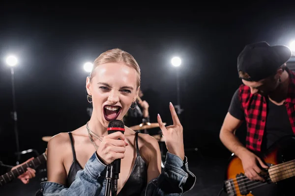 КИЕВ, УКРАИНА - 25 августа 2020 года: Блондинка с рок-жестом поет в микрофоне во время репетиции рок-группы на размытом фоне — стоковое фото