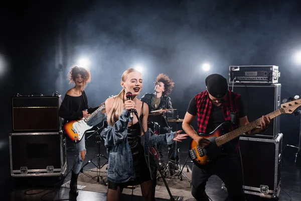 KYIV, UKRAINE - 25 de agosto de 2020: Mulher de olhos fechados cantando enquanto segura rack de microfone perto de músicos da banda de rock em fundo embaçado — Fotografia de Stock