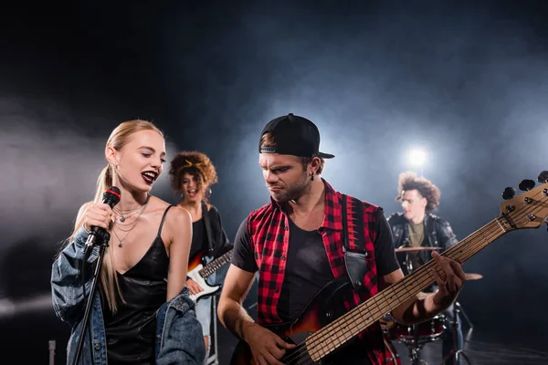 KYIV, UKRAINE - AUGUST 25, 2020: Smiling blonde woman singing near guitarist during rock band rehearsal with backlit on blurred background — Stock Photo