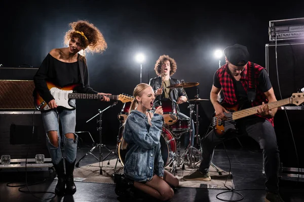 KYIV, UKRAINE - 25 AOÛT 2020 : Femme blonde avec microphone hurlant alors qu'elle était assise à genoux pendant la performance du groupe de rock avec rétroéclairage sur noir — Photo de stock