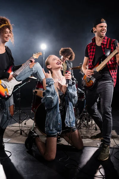 KYIV, UCRANIA - 25 de agosto de 2020: mujer con micrófono mostrando un cartel de rock sentada de rodillas cerca de músicos con retroiluminación sobre fondo negro - foto de stock