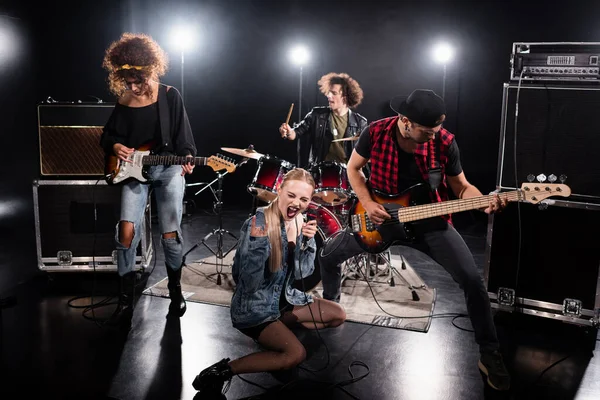 KYIV, UKRAINE - AGOSTO 25, 2020: mulher agachada e mostrando sinal de rock, enquanto gritava em microfone durante o ensaio da banda de rock com retroiluminação em preto — Fotografia de Stock