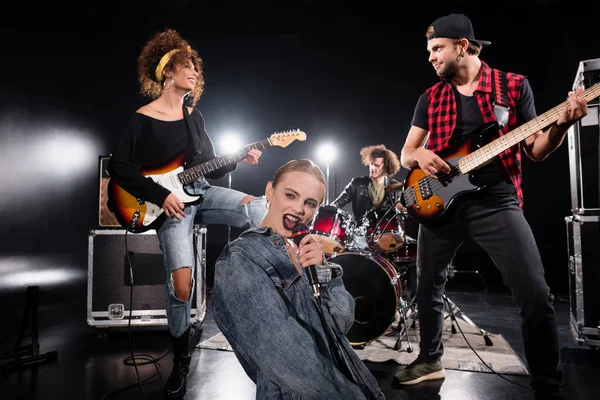 KYIV, UCRANIA - 25 de agosto de 2020: Vocalista femenina cantando en micrófono mientras está sentada cerca de guitarristas con batería retroiluminada y borrosa en el fondo - foto de stock