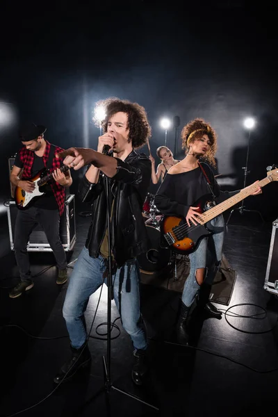 KYIV, UKRAINE - AUGUST 25, 2020: Curly vocalist pointing with finger while singing near guitarists with backlit and female drummer on background — Stock Photo