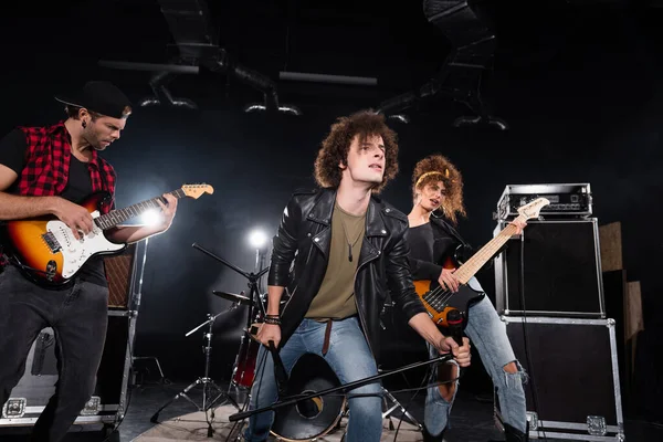 KYIV, UKRAINE - AUGUST 25, 2020: Curly vocalist looking away while holding microphone rack, standing near guitarists with backlit on black — Stock Photo