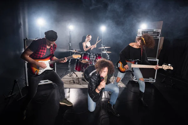 KYIV, UKRAINE - AUGUST 25, 2020: Curly vocalist looking away while sitting on knee during rock band performance with backlit on black — Stock Photo