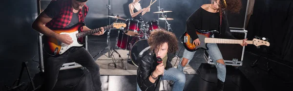 KYIV, UKRAINE - AUGUST 25, 2020: Curly vocalist looking away while sitting on knee during rock band performance, banner — Stock Photo