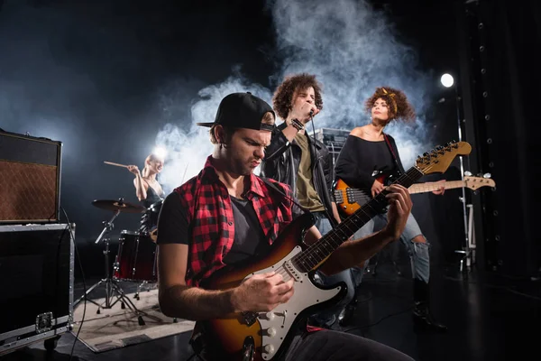 KYIV, UKRAINE - 25 de agosto de 2020: Músico de la banda de rock tocando el bajo sentado cerca del vocalista rizado y guitarrista con humo y batería femenina en el fondo - foto de stock