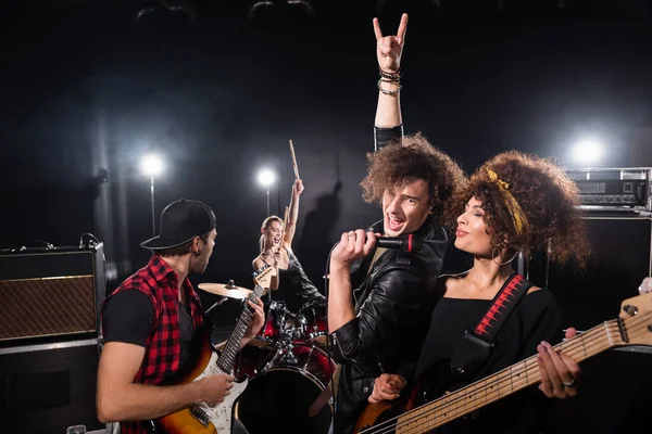 KYIV, UCRANIA - 25 de agosto de 2020: Emocionado vocalista con gesto de rock sosteniendo micrófono cerca de una mujer con guitarra eléctrica durante la actuación de la banda de rock en negro - foto de stock