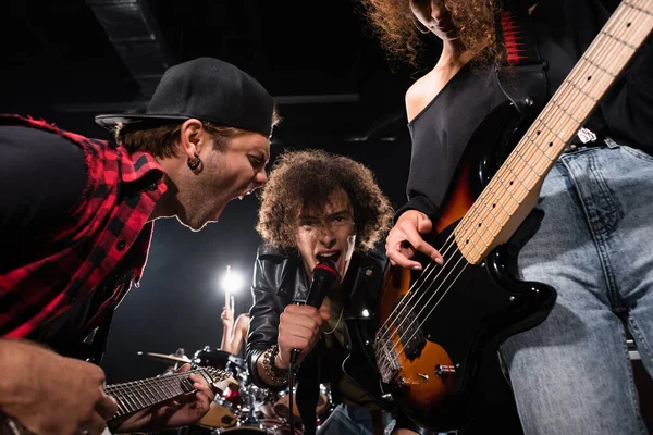 KYIV, UCRANIA - 25 de agosto de 2020: Vista de bajo ángulo del vocalista rizado mirando a la cámara mientras grita con el guitarrista con retroiluminación sobre fondo borroso - foto de stock