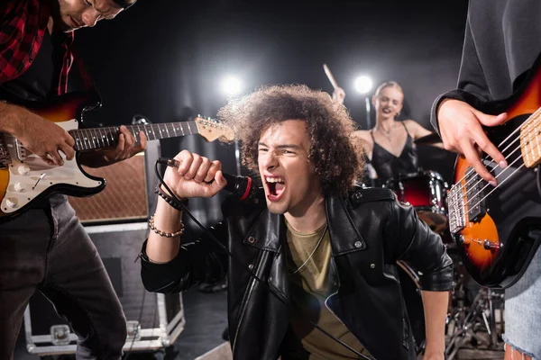 KYIV, UKRAINE - AGOSTO 25, 2020: Vocalista encaracolado gritando em microfone enquanto se senta perto de guitarristas com o baterista feminino retroiluminado e borrado no fundo — Fotografia de Stock