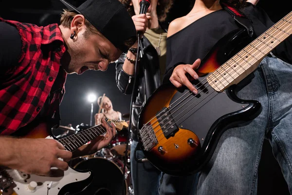 KYIV, UCRANIA - 25 de agosto de 2020: Guitarrista gritando mientras se inclina hacia adelante cerca de músicos de bandas de rock con retroiluminación sobre un fondo borroso - foto de stock