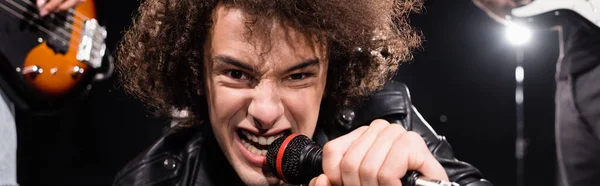 KYIV, UKRAINE - AUGUST 25, 2020: Angry vocalist with microphone signing while looking at camera with backlit and blurred guitarists on black, banner — Stock Photo