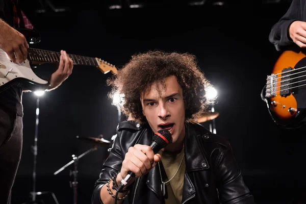 KYIV, UKRAINE - AUGUST 25, 2020: Curly vocalist with microphone looking at camera near musicians with electric guitars on blurred background — Stock Photo