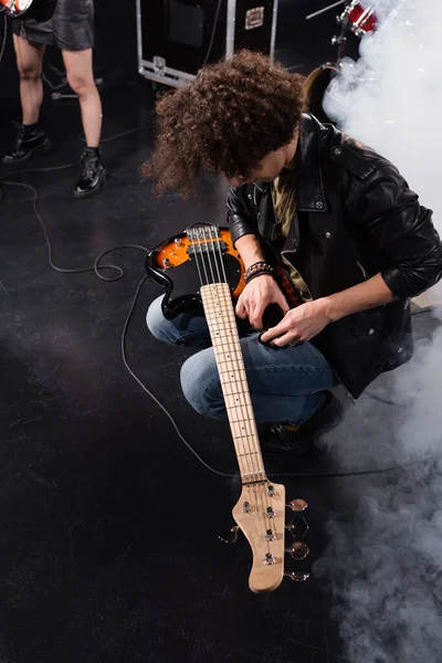 KYIV, UKRAINE - AUGUST 25, 2020: Overhead view of curly musician squatting with electric guitar on knees during rock band rehearsal — Stock Photo
