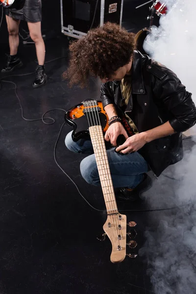 KYIV, UCRANIA - 25 de agosto de 2020: Vista aérea del músico rizado en cuclillas con guitarra eléctrica sobre las rodillas durante el ensayo de la banda de rock - foto de stock