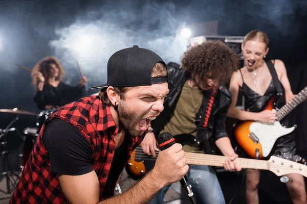 KYIV, UCRANIA - 25 de agosto de 2020: Vocalista gritando en micrófono mientras se inclina hacia adelante cerca de músicos de bandas de rock sobre un fondo borroso - foto de stock