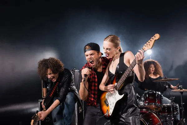 KYIV, UKRAINE - AUGUST 25, 2020: Vocalist shouting in microphone standing near guitarists with woman playing drums and backlight on black — Stock Photo