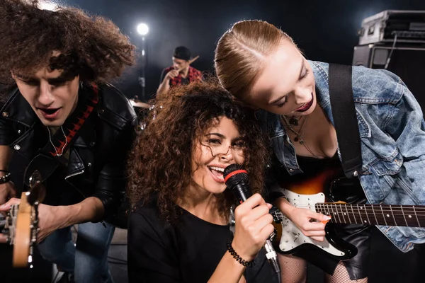 KIEW, UKRAINE - 25. AUGUST 2020: Lockige Frau mit Mikrofon blickt in die Kamera in der Nähe von Rockband-Gitarristen mit hinterleuchtetem und verschwommenem Schlagzeuger im Hintergrund — Stockfoto