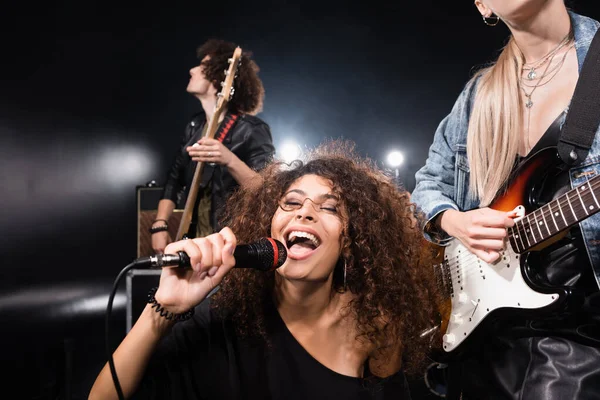 KYIV, UCRANIA - 25 de agosto de 2020: Mujer rizada feliz con micrófono cantando cerca de guitarristas de bandas de rock con luz de fondo en negro - foto de stock