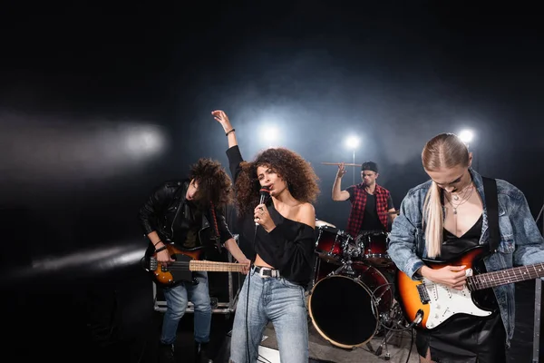 KYIV, UKRAINE - AUGUST 25, 2020: Curly woman with hand in air singing standing near guitarists and drummer with backlit on black — Stock Photo