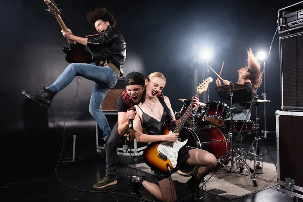 KYIV, UKRAINE - AUGUST 25, 2020: Vocalist shouting in microphone while leaning forward near guitarist jumping up and woman sitting on knee with backlit on black — Stock Photo