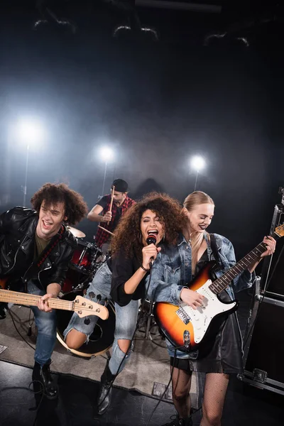 KYIV, UKRAINE - 25 de agosto de 2020: Vocalista rizada cantando y abrazando a la guitarrista femenina durante el ensayo de la banda de rock con retroiluminación en negro - foto de stock