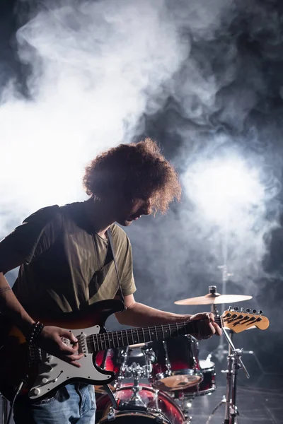 KYIV, UKRAINE - 25 de agosto de 2020: músico de banda de rock tocando guitarra com pick near drum kit com fumaça no fundo — Fotografia de Stock