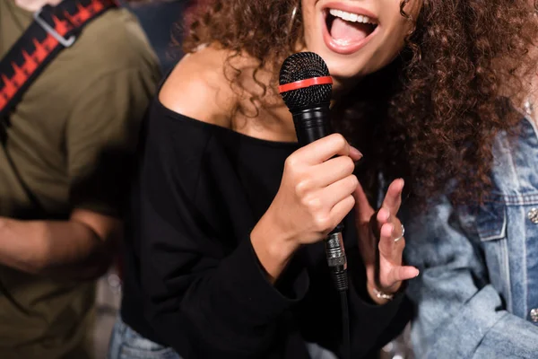 Обрезанный вид кудрявой вокалистки с микрофоном, поющей рядом с музыкантами рок-группы на размытом фоне — стоковое фото