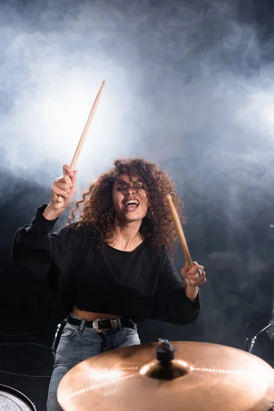 Baterista femenina con los ojos cerrados gritando, mientras toca en la placa de tambor con humo sobre fondo negro - foto de stock