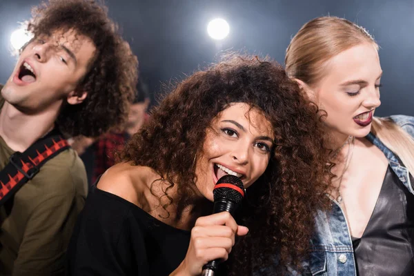 Vocalista de banda de rock feliz con micrófono cantando cerca de músicos con retroiluminación sobre fondo borroso - foto de stock