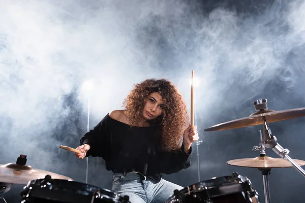 Female musician with drumsticks playing on drum kit while looking at camera with smoke on background — Stock Photo